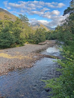 A tranquil river flows through a wooded landscape with mountains in the background and a clear blue