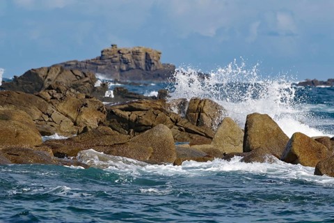 Waves crashing against rocks with a rocky background under a blue sky, Isles of Scilly, Cornwall,