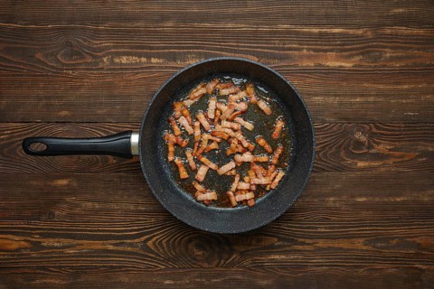 Sizzling strips of bacon fry in a black skillet, releasing savoury aroma and fat on rustic wooden