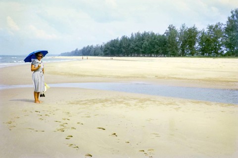 British woman holding umbrella standing on sandy beach south of estuary, Rompin, East coast,