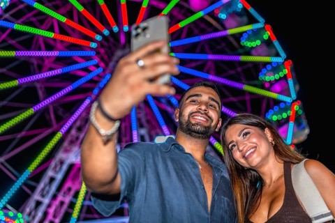 Young couple smiling and taking a selfie with smartphone in front of a colorful ferris wheel at an