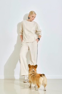 A woman dressed in light, casual attire stands against a plain wall while a corgi dog curiously