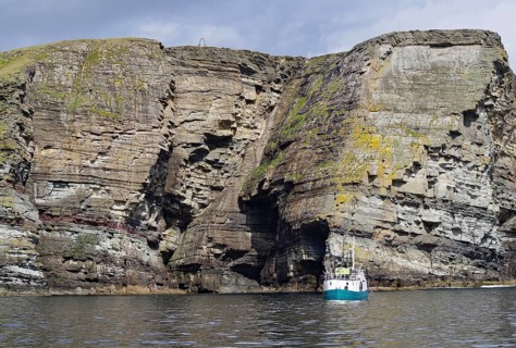 Boat in front of massive cliffs in the sea under a cloudy sky, Noss, Shetland Islands, Scotland,