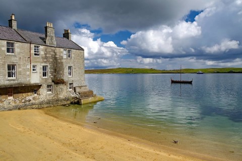 Old stone cottage on quiet loch shore with sandy beach and passing boat under cloudy sky, Murder in