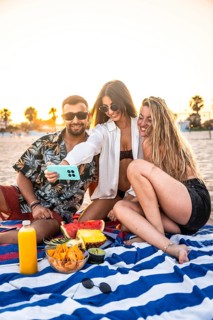 Three friends are taking a selfie with smartphone during a picnic at the beach, enjoying snacks and
