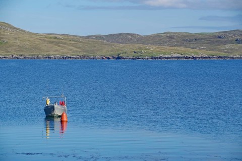 Lonely boat next to buoy on calm water, hilly landscape behind under clear sky, Hillswick,