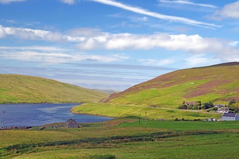 Green hilly landscape with a river and scattered houses under a blue, slightly cloudy sky, Voe,