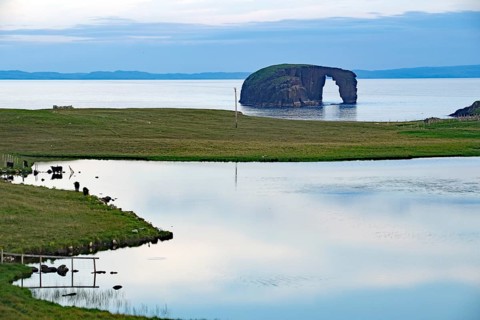 Calm lake with grassy shores and a rock formation on the horizon, dore holm Dore Holm, Hillswick,