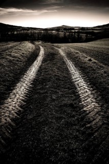 Tire tracks cut through a dark, rural field, leading toward rolling hills under a moody sky at dusk