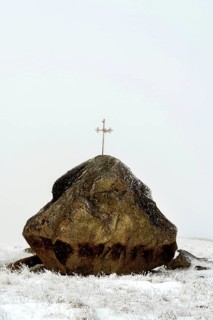 A large rock topped with a cross stands starkly against the winter landscape of the Aubrac Plateau