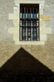 A barred window positioned on a rough stone wall casts a strong shadow below. Sunlight enhances the