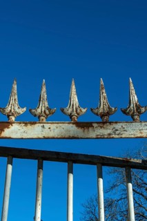A weathered wrought iron fence stands tall under a bright blue sky. The fence has pointed spikes,