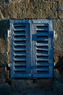 Sunlight casts shadows on a pair of weathered blue shutters attached to an old stone wall. The