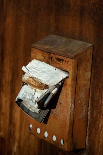A rusty mailbox attached to a weathered wall is bursting with various papers and ads, indicating
