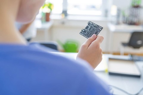Person holding a circuit board in front of a blurred background with a window view