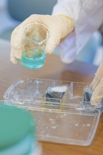 Person in gloves holding a beaker with blue liquid over a tub, Battery Research, Nagold, Germany