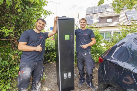 Two technicians pose proudly next to a closed power box outdoors, installing a charging station for