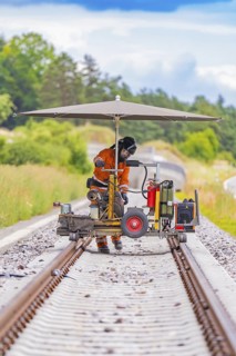Worker using a machine on railway tracks under an umbrella, surrounded by greenery, grinding work