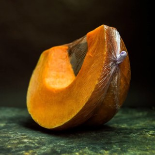 A vibrant slice of pumpkin is displayed, encased in plastic wrap. The dark background enhances the
