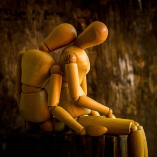 A wooden mannequin is positioned on a rough wooden surface, looking contemplative against a