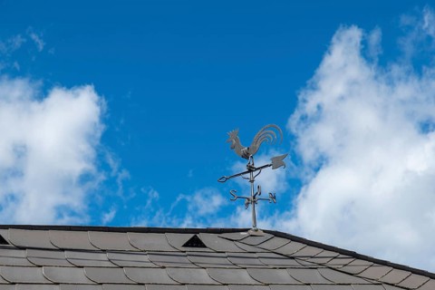 A metal weathercock on the roof in front of a blue sky with clouds