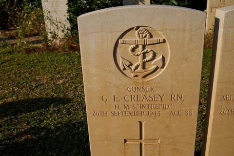 A maritime emblem on a gravestone with personal information, surrounded by grass and shadow,