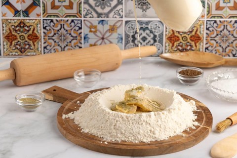 Milk is poured into a well of flour on a wooden board, surrounded by ingredients
