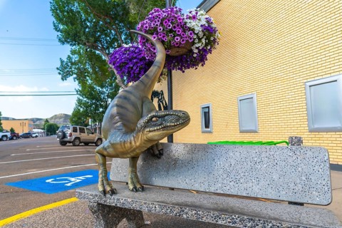 Dinosaur statue resting on a bench in front of a brick building, surrounded by flower baskets in
