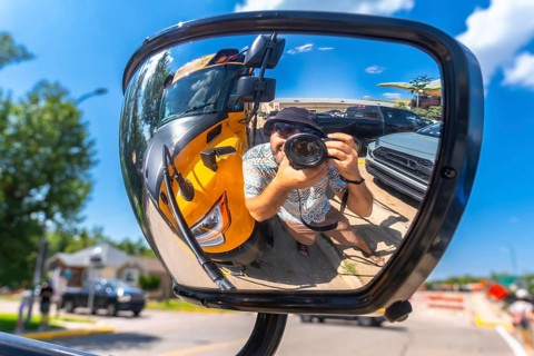 Photographer is taking a selfie reflected in the mirror of a truck, with a yellow school bus and