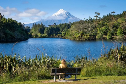 A woman sitting on a park bench at Lake Mangamahoe with views of Mount Taranaki. Egmont National
