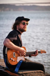 Hipster street musician in black playing electric guitar in the street sitting on pier embankment