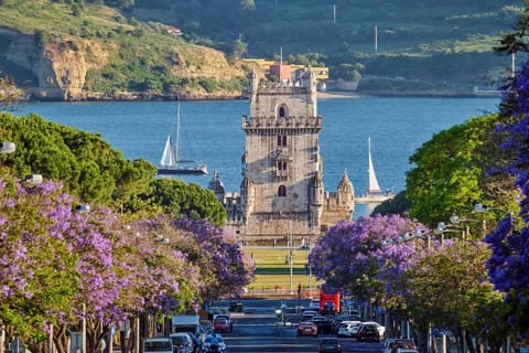 Scenic view of Belem Tower surrounded by blooming jacaranda trees, with sailboats on the Tagus