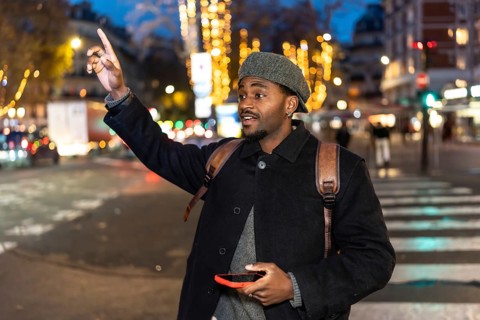Young man in a winter coat stands on a busy city street at night, holding a smartphone and raising