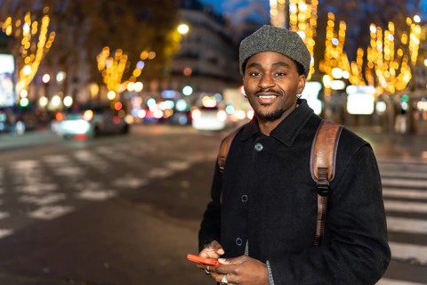 Young black man smiling in an urban setting at night, holding a mobile phone and standing on a city