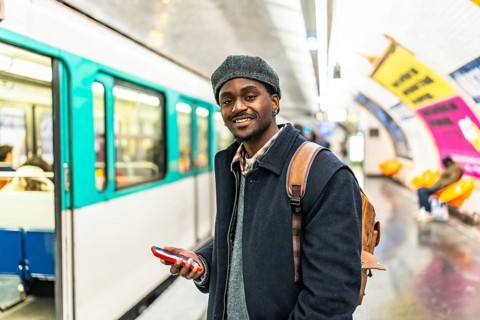 Young black man in a beret smiles at camera while holding a phone and backpack on a paris subway