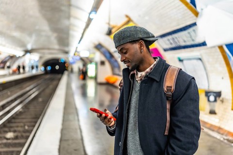 Young black man in a flat cap and backpack checks his smartphone while waiting on a busy urban