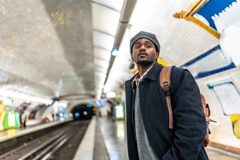 Young black man in a beret with a backpack stands on a tiled paris metro platform, looking up while