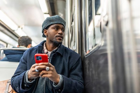 Young black man seated on subway, dressed for winter, holding smartphone and gazing out the window