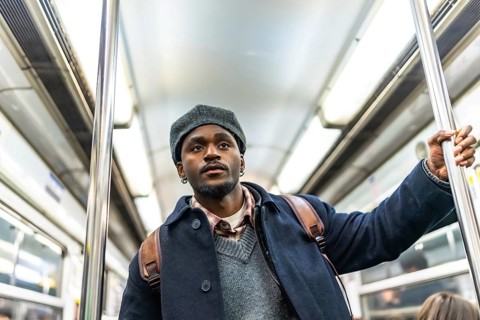 Young black man in winter coat and hat stands holding a subway pole on a crowded metro, looking