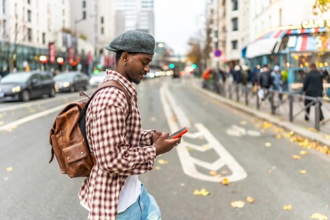Young man wearing a beret and backpack walking across a city street, looking down at his smartphone