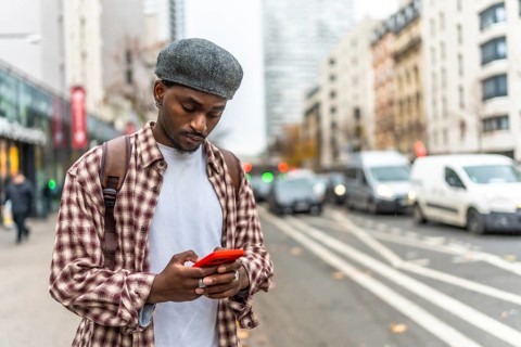 Young black man standing on an urban street, wearing a plaid shirt and a cap, engrossed in using