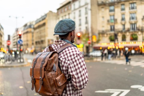 Young man wearing a cap and brown backpack stands on the curb of an urban street, looking across