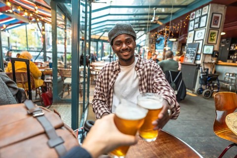 Young man smiling and clinking beer glasses with a friend in a casual pub, celebrating friendship,