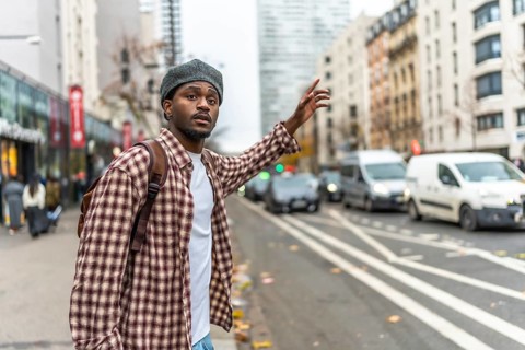 Young black man wearing a beret and a plaid shirt standing on an urban sidewalk, raising his hand