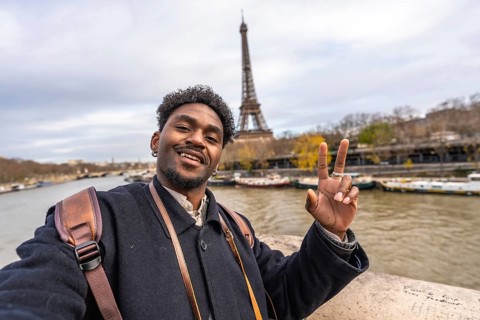 Smiling young man vlogging a selfie on a paris trip, flashing a peace sign with the eiffel tower