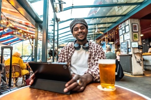 Young black man in a plaid shirt and hat, smiling at camera while enjoying a beer and using a