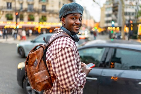 Young black man smiling, wearing a beret and backpack, holding a smartphone while walking on a busy