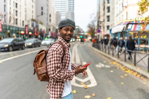 Young black man in a checkered shirt and backpack smiles at the camera while checking his phone on