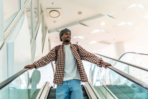 Young black man riding a modern escalator in a downtown shopping mall, wearing a backpack and