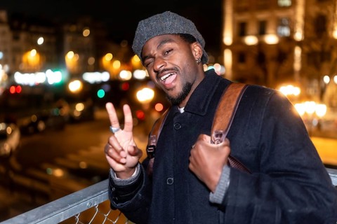 Young man in a flat cap standing outdoors in the city at night, winking at the camera and showing a